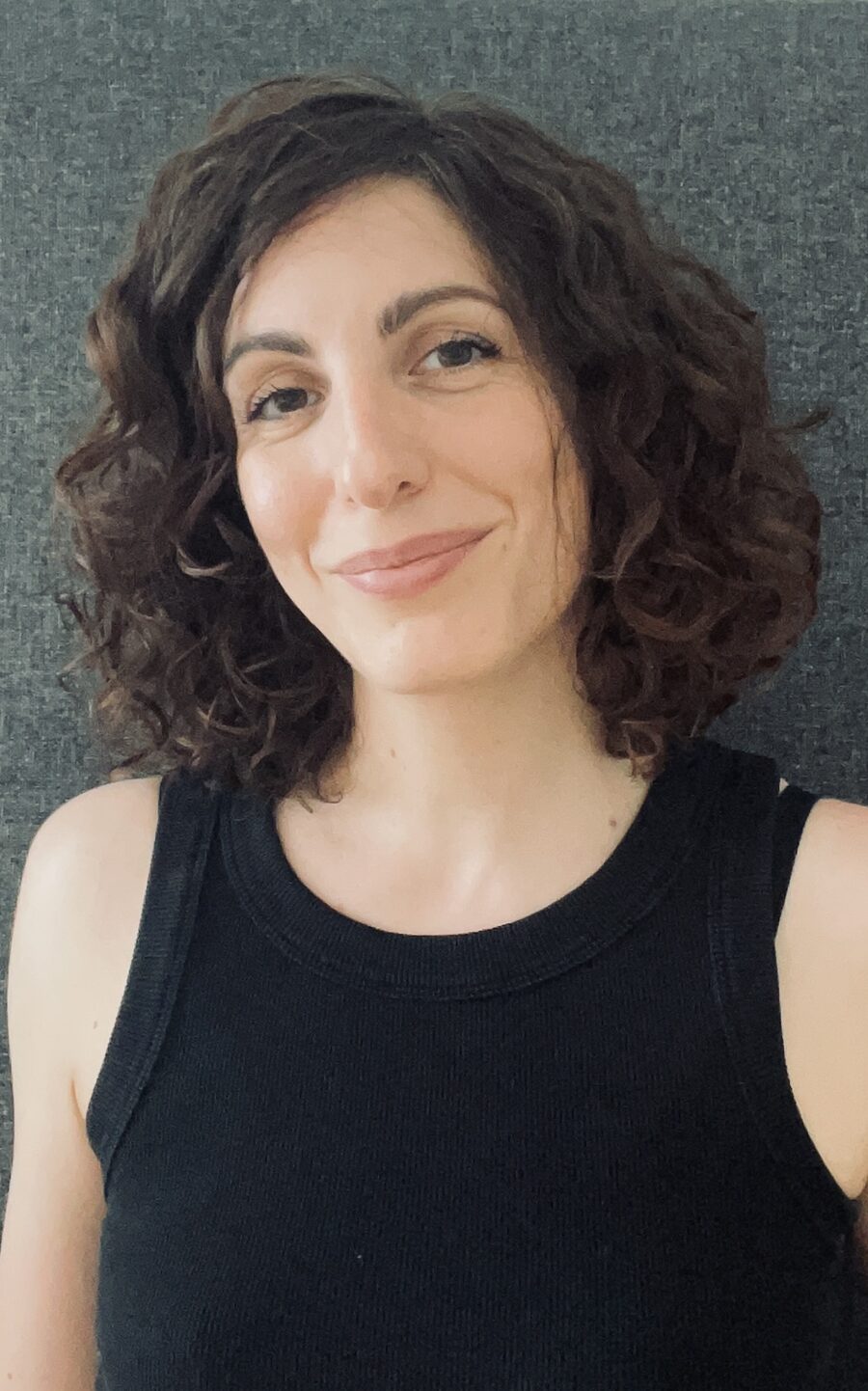 Portrait of Marie Kolkenbrock, smiling: a white woman with shoulder-length dark curly hair, against a grey wall