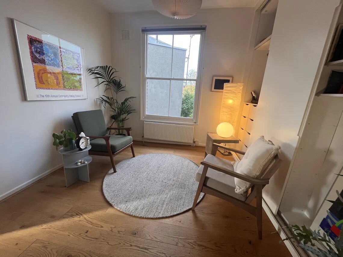 The therapy room in Finsbury Park: two armchairs facing each other across a round grey rug, a tall paper floor lamp and a glowing globe lamp, a large plant beside a sash window with trees outside, and a framed print on the wall